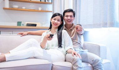 Kanepede birlikte televizyon seyreden gülümseyen bir çift kadın uzaktan kumandayı tutarken erkek işaret eder.