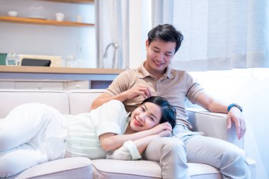 Mutlu bir çift, parlak bir evde, kanepede dinlenirken kadın başını erkeğin kucağına koyuyor.