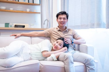 Gülümseyen bir çift, parlak, modern bir oturma odasındaki bir kanepede rahatlamış ve boş zamanlarını değerlendiriyor.