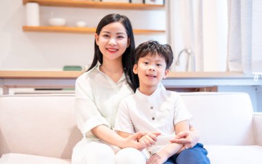 Neşeli bir Asyalı anne ve oğlu modern ışık dolu bir odada parlak bir kanepede gülümsüyor.