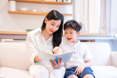 Bir kadın, genç bir çocuğa kitap okuyor. İyi aydınlatılmış, modern görünümlü bir evde, bir kanepede otururken.