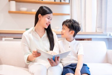 Mutlu bir anne evde oğluna kitap okur. Her ikisi de sıcak bir yuva ortamında sıcak bir gülümseme paylaşır.