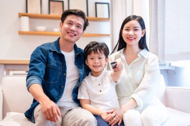 Gülümseyen bir aile birlikte kanepede oturuyor. Oğlu televizyon izlerken uzaktan kumandayı işaret ediyor.