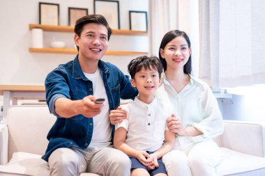 Üç kişilik gülümseyen aile, parlak bir oturma odasında kanepede oturup televizyon izliyorlar.