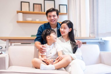 Üç kişilik bir aile, krem renkli bir koltukta otururken ebeveynler oğullarına sarılıyorlar. Hepsi gülümsüyor.