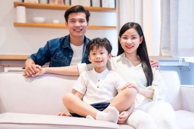 Üç kişilik gülümseyen bir aile modern ışık dolu oturma odalarında birlikte otururlar.