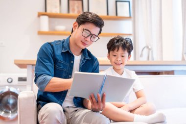 Bir baba ve oğlu, modern minimalist evlerinde bir kanepede birlikte kitap okuyorlar.