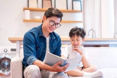 Bir adam, parlak bir oturma odasında bir çocuğa kitap okuyor ve okuma sevgisini besliyor.