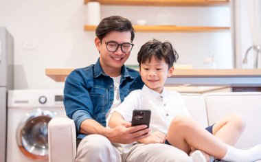 Gülümseyen bir baba ve oğul kanepede yan yana otururken telefonla içeriğe bakarlar.