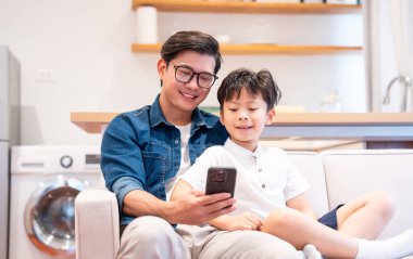 Bir baba ve oğul oturma odasındaki koltukta cep telefonuna bakarken gülüşürler.