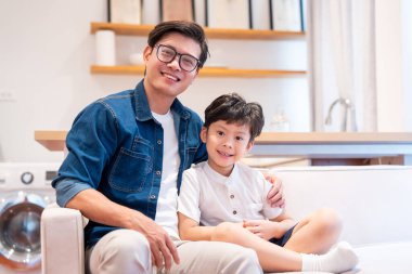 Gözlüklü bir baba ve oğlu içeride bir koltukta otururken ve gülümserken görülüyor.