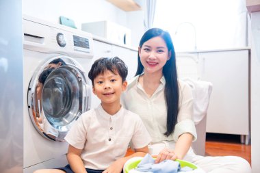 Gülümseyen bir anne ve oğlu, aydınlık ve modern görünümlü bir çamaşırhanede birlikte çamaşır yıkıyorlar.