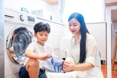 Bir anne ve oğlu birlikte çalışarak çamaşır katlıyor. Parlak bir odada, ön yükleme makinesinin yanında.