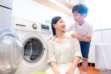 Bir anne ve oğlu çamaşır odasında birlikte çamaşır yıkıyorlar. Aydınlık bir odada.