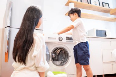 Bir çocuk, anne ve babasının aydınlık, modern bir mutfakta izlediği gibi çamaşırları çamaşır makinesine dolduruyor.