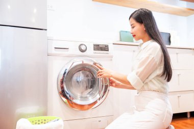 Beyazlı genç bir kadın temiz, modern bir mutfakta çamaşır makinesine çamaşır yüklüyor.