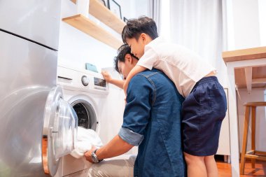 Oğlu sırtına sevgiyle sarılırken bir baba çamaşır makinesine çamaşır doldurur.