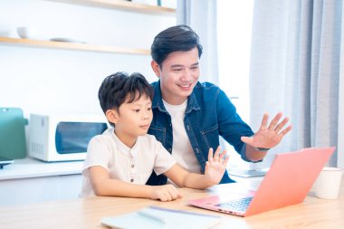 Bir baba ve oğlu dizüstü bilgisayar kullanıyor gülümsüyor ve ışık dolu bir mutfak alanına el sallıyor.