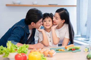 Aile mutfakta birlikte sağlıklı bir yemek hazırlıyor. Ebeveynler oğullarını yanaklarından öpüyor.