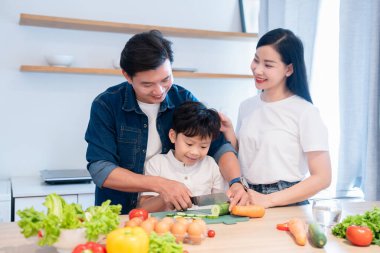 Mutfakta bir aile var. Baba oğluna sebze doğramasında yardım ediyor ve annesi yakınlarda onu izliyor.