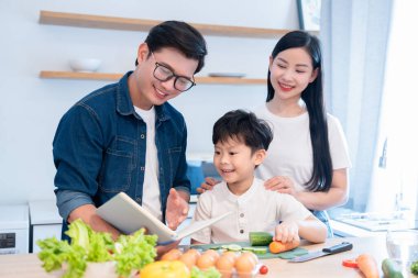 Neşeli Asyalı aile mutfakta birlikte yemek pişiriyor. Tablete bakarak sağlıklı bir yemek hazırlıyorlar.