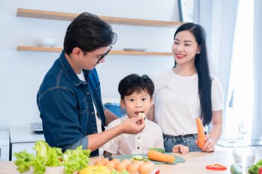 Parlak bir mutfakta bir aile. Baba oğlunu bir sebzeyle besliyor. Anne de elinde havuçla izliyor.