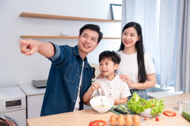 Mutfakta üç kişilik bir aile kahvaltı hazırlar. Baba pencereyi gösterir ve oğlu yumurtaları karıştırır.