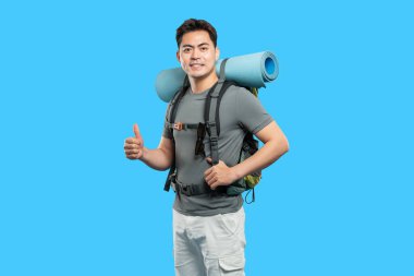 Sırt çantası ve uyku paspası olan gülümseyen Asyalı adam açık mavi arka planda başparmak kaldırıyor.