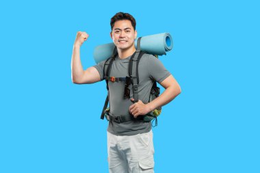 Sırt çantası takan ve uyku paspası taşıyan bir yürüyüşçü mavi arka planda kas gücünü gösterir.