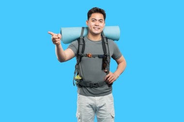 Sırt çantalı ve seyahat minderli gülümseyen Asyalı yürüyüşçü gideceği yeri gösteren mavi bir yolda duruyor.