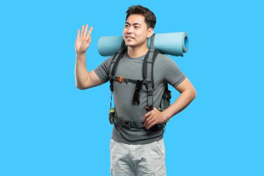 Sırt çantalı genç adam açık mavi bir arkaplanda duruyor. Maceraya hazır bir dost dalgası sunuyor.