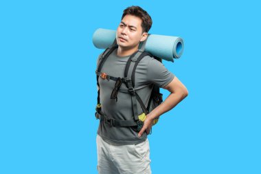 Yürüyüş yapan ve sırt ağrısı çeken bir adam mavi arka planda sırt çantası ve uyku paspası takıyor.