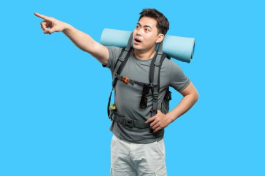 Sırt çantası ve uyku paspası ile coşkulu bir erkek yürüyüşçü heyecanlı bir şekilde mavi bir arkaplanı işaret ediyor.