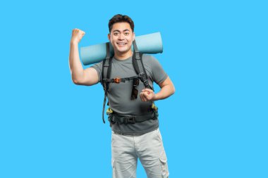 Hevesli genç adam yürüyüş malzemeleriyle poz veriyor. Sırt çantası takıyor ve mavi bir arka fonda hasırı katlıyor.