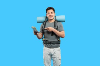 Sırt çantası ve uyku tulumuyla heyecanlı bir yürüyüşçü bir harita tutar ve parlak mavi arka planda gülümser.