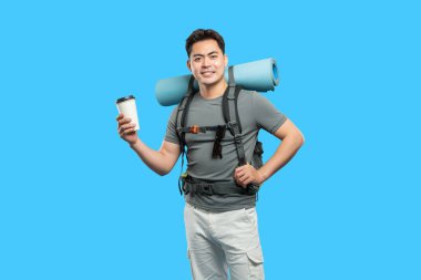 Sırt çantalı gülümseyen adam dışarıda yürüyüşe hazır mavi bir fonda kahve tutuyor.