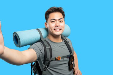 Mavi arka planda sırt çantası ve yoga minderi takan dostane bir gülümsemesi olan genç bir gezgin.