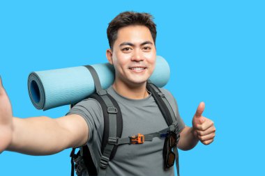 Sırt çantası ve uyku tulumu taşıyan neşeli bir yürüyüşçü başparmağıyla coşkulu bir selfie çeker.