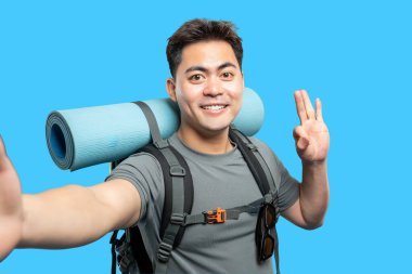 Gülümseyen gezgin sırt çantasıyla selfie çekiyor ve mavi arka plana barış işareti koyuyor.