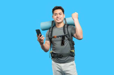 Mavi arka planda sırt çantalı Asyalı adam telefonunu kontrol ediyor. Gülümsüyor ve maceraya hazır.