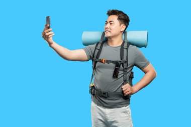 Sırt çantalı bir adam mavi arka planda seyahat ve teknoloji gösteren bir selfie çekiyor.