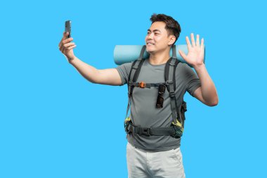 Neşeli adam selfie çekerken mavi bir sırt çantasına ve yürüyüş takımına el sallıyor.