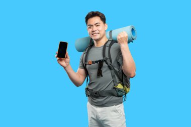 Gülümseyen Asyalı yürüyüşçü, telefonun mavi arka planda olduğunu gösteriyor. Sırt çantası, güneş gözlüğü uyku paspası da dahil.