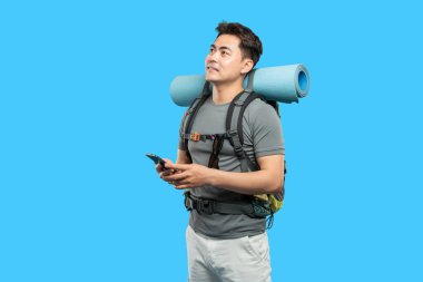 Düşünceli bir Asyalı sırt çantası parlak mavi bir stüdyo arka planında ekipman taşırken akıllı bir telefon tutar.