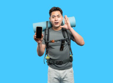 Hevesli genç yürüyüşçü mavi arka planda boş akıllı telefon çantası ve uyku paspası sergiliyor.