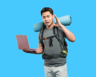 Sırt çantası ve uyku tulumu olan bir adam bir sonraki macerasını planlamak için mavi arka planda dizüstü bilgisayar kullanıyor.