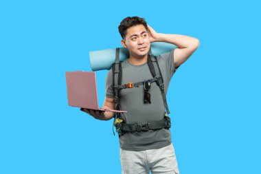 Sırt çantası ve dizüstü bilgisayarı olan bir adam mavi bir arkaplanın önünde kafasını düşünceyle kaşıyarak duruyor.