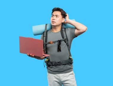 Sırt çantası ve uyku tulumuyla şaşkın bir yürüyüşçü mavi bir arkaplana karşı dizüstü bilgisayar tutuyor.