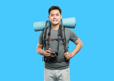 Sırt çantası ve kamerası olan bir adam açık hava macerasına hazır mavi bir arka planda duruyor.