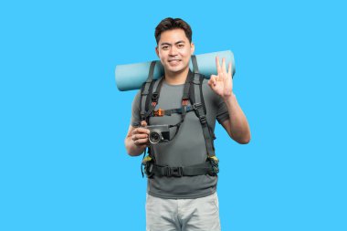 Mavi ekranın önünde poz veren bir turist sırt çantası takıyor, tamam işareti veriyor ve elinde bir kamera tutuyor.